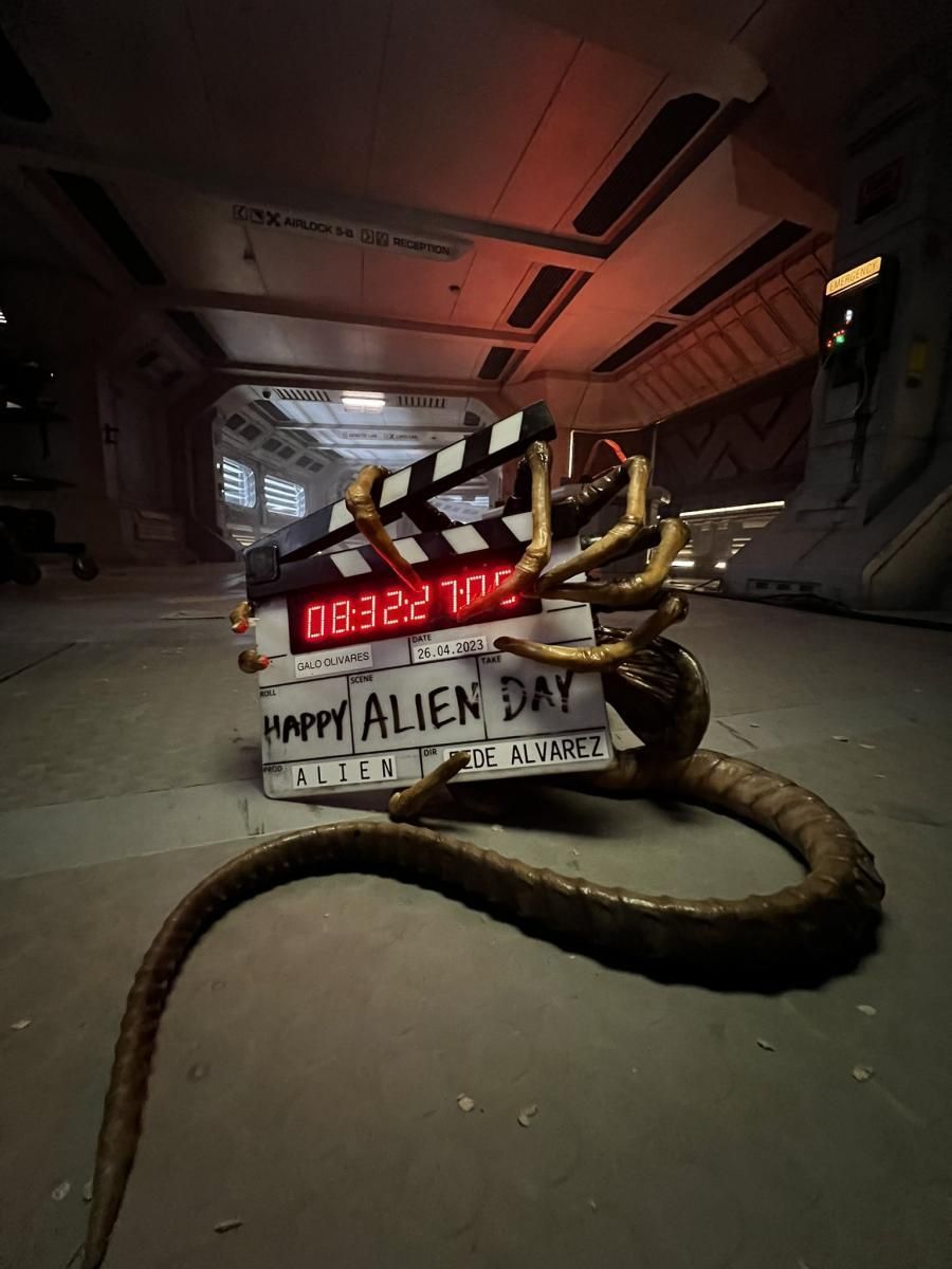 Una claqueta de película con la inscripción 'Happy Alien Day' apoyada en el suelo de una nave espacial con un ambiente oscuro y claustrofóbico. La claqueta está enredada con la cola característica de un xenomorfo de la franquicia 'Alien', sugiriendo una escena de la película 'ALIEN' dirigida por Fede Alvarez, ambientada para ser filmada.