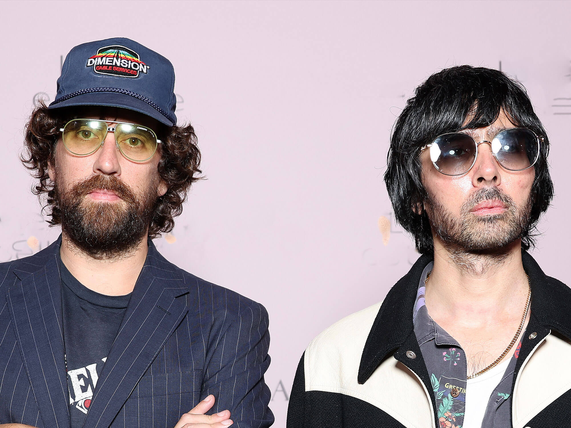 Dos hombres con apariencia de músicos posan para una fotografía. El de la izquierda lleva una gorra azul con visera, gafas claras y una barba completa, vistiendo un traje a rayas sobre una camiseta con estampado. El de la derecha lleva gafas de sol redondas de tono oscuro y una chaqueta color crema con detalles negros sobre una camisa estampada, con una expresión seria.