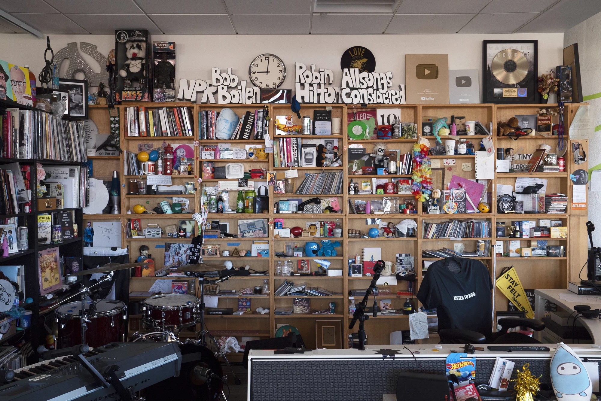 La imagen muestra un espacio de trabajo densamente decorado, presumiblemente para Tiny Desk Concerts de NPR Music, como se sugiere por las letras en la pared. El lugar está lleno de objetos variados: libros, discos, figuritas, instrumentos musicales como teclados y una batería, y equipo de grabación. Hay placas de YouTube, marcos con discos, y arte variado. Un sello distintivo de la serie es el ambiente personal y caótico que se refleja en el conjunto de objetos y recuerdos, que otorgan un aire de creatividad y pasión por la música.