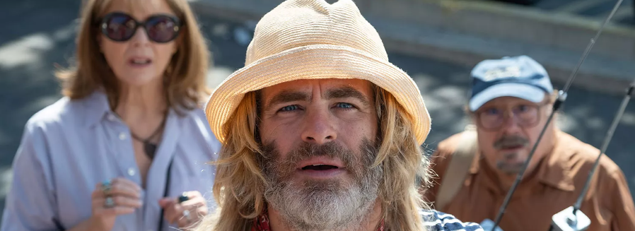 La imagen muestra a tres personas, posiblemente actores, en lo que parece ser una escena de una película o serie. En primer plano, hay un hombre con una barba rubia y pelo largo, lleva una camiseta a rayas y una gorra de pescador anaranjada. Tiene una expresión seria o pensativa. Detrás de él, una mujer con gafas de sol y camisa azul claro está hablando y parece estar en movimiento. Al lado de ella, hay otro hombre mayor con una gorra de beisbol y camisa color canela, sosteniendo una caña de pescar y mirando hacia el mismo lugar que el hombre en primer plano. El ambiente sugiere una actividad al aire libre, quizás relacionada con la pesca o un viaje de ocio.